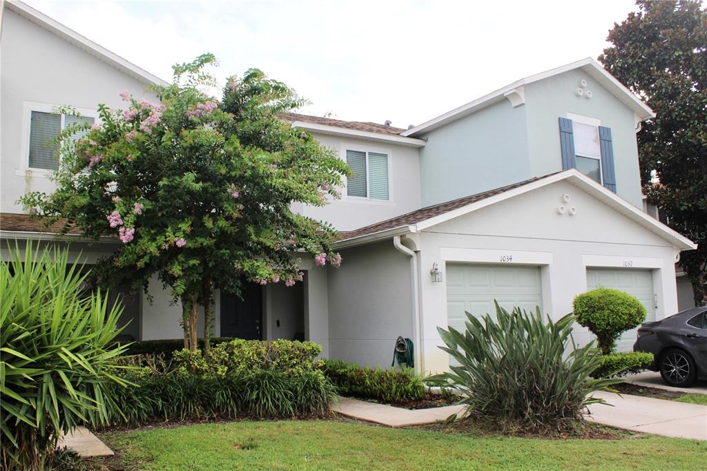 A two-story townhouse with a light gray exterior, two garage doors, and a flowering tree — 1034 CHALCEDONY STREET, KISSIMMEE, FL