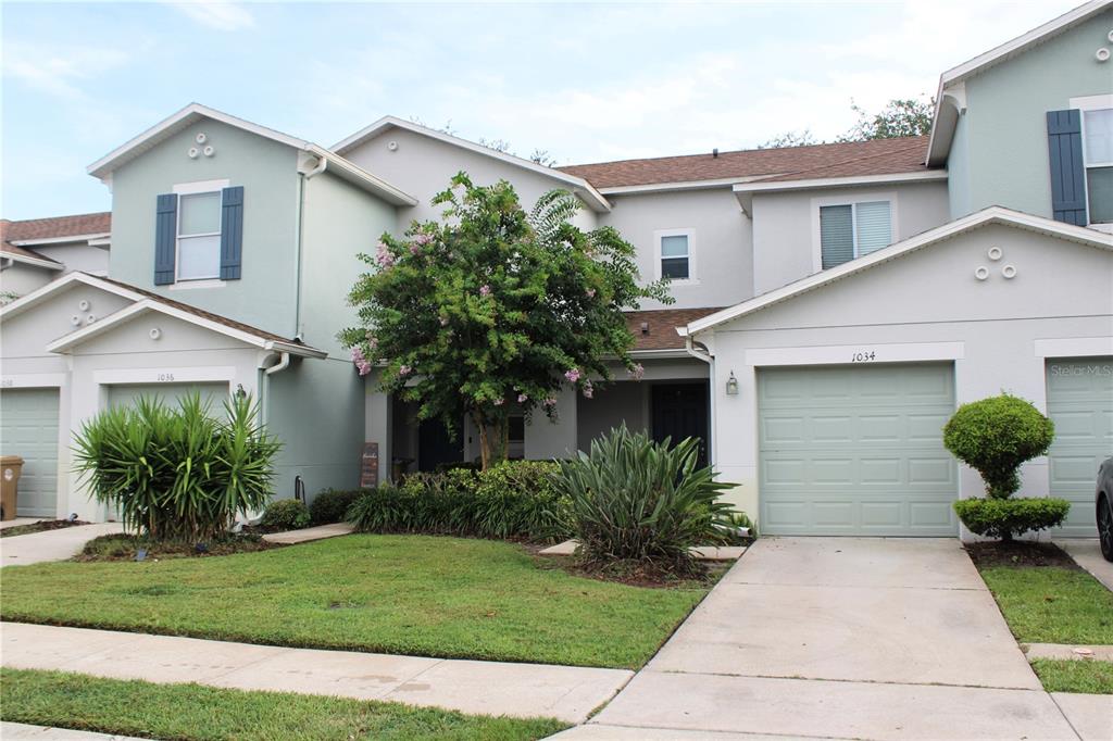 A two-story townhouse exterior features light-colored stucco, a green garage door, and a front lawn — 1034 CHALCEDONY STREET, KISSIMMEE, FL