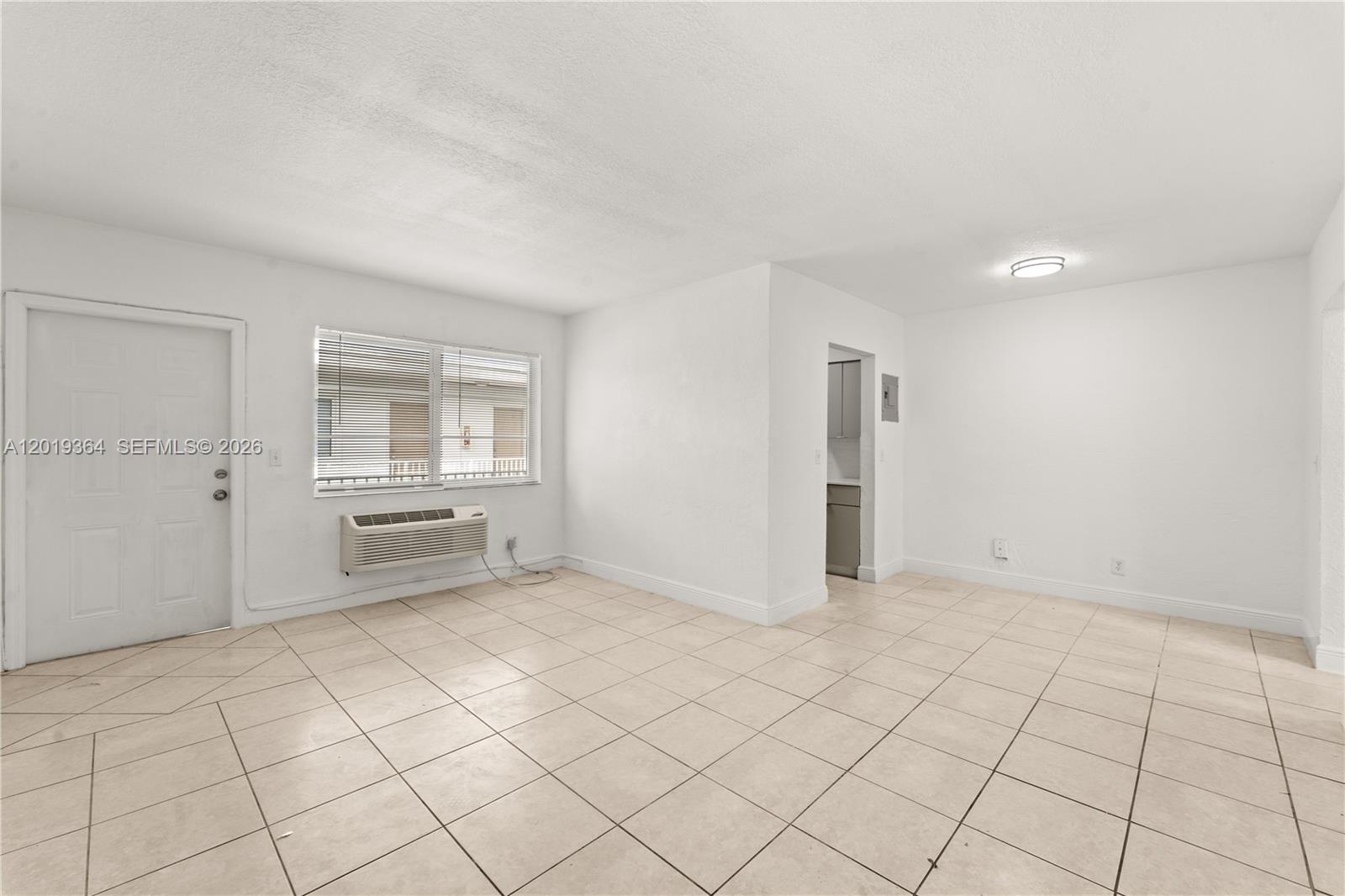 A bright, empty room features white walls, light-colored tile flooring, a window, and an air-conditioning unit — 6905 Rue Vendome # 8, Miami Beach FL 33141, MIAMI BEACH, FL