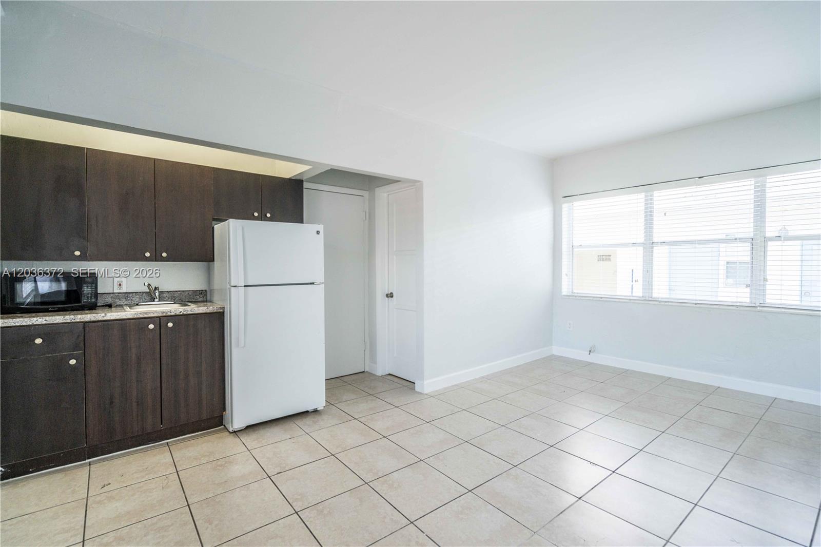 An empty room features white walls, a large window with blinds, and light-colored tile flooring — 8315 Byron Ave # 7A, Miami Beach FL 33141, MIAMI BEACH, FL