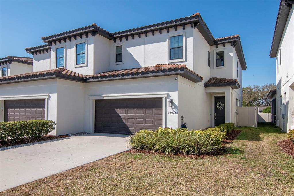 A two-story stucco townhouse features a brown garage door, tiled roof, and front landscaping — 13022 SANCTUARY VILLAGE LANE, TAMPA, FL