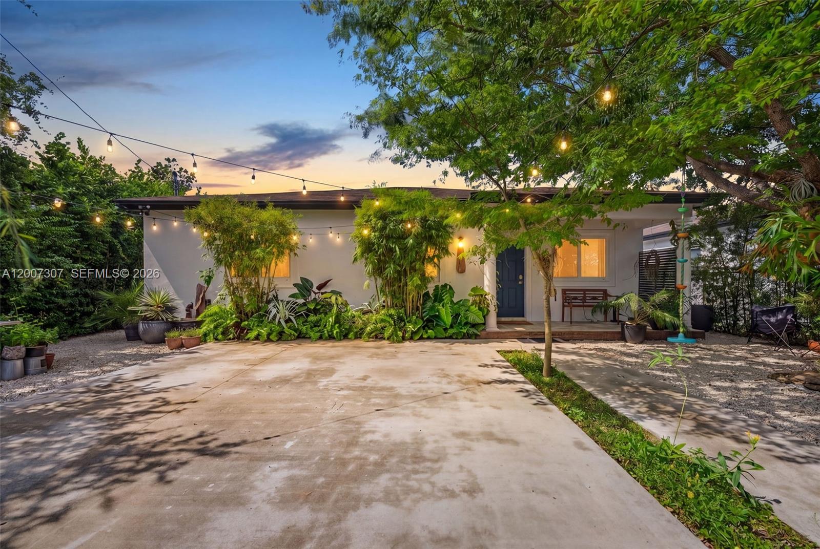 A single-story house with a concrete driveway, string lights, and lush landscaping at dusk — 12310 SW 216th St, Miami FL 33170, MIAMI, FL