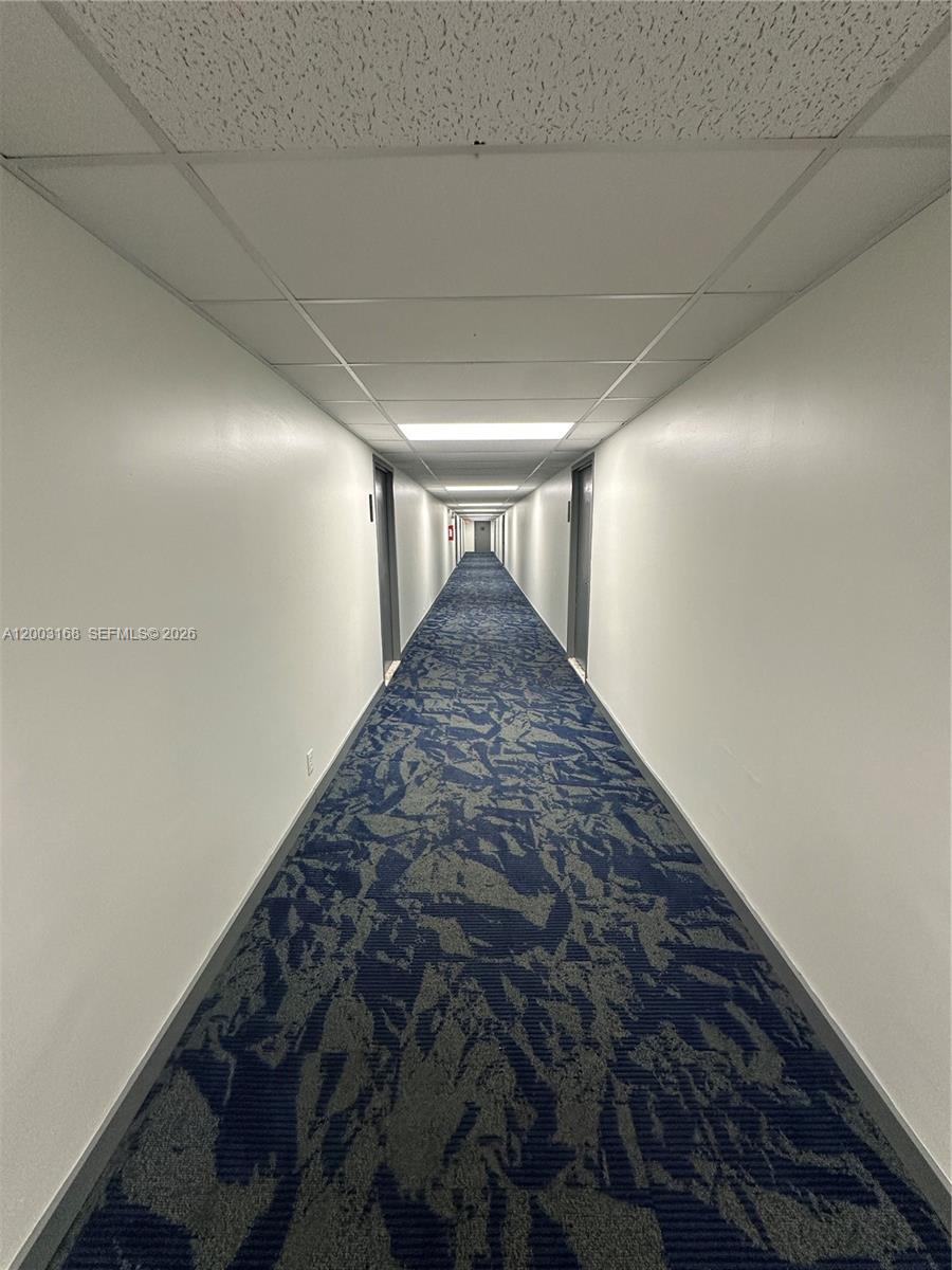 A long, narrow hallway features white walls, a drop ceiling, and patterned blue carpet — 701 Three Islands Blvd # 110, Hallandale Beach FL 33009, HALLANDALE BEACH, FL