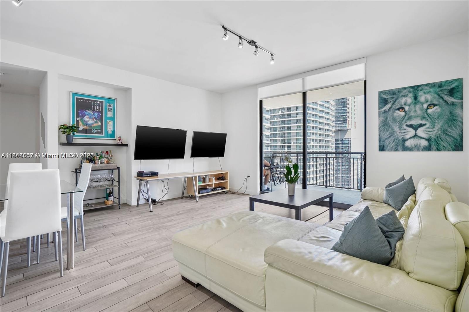 A living room features a white sectional sofa, two wall-mounted televisions, and a balcony view — 999 SW 1st Ave # 2717, Miami FL 33130, MIAMI, FL