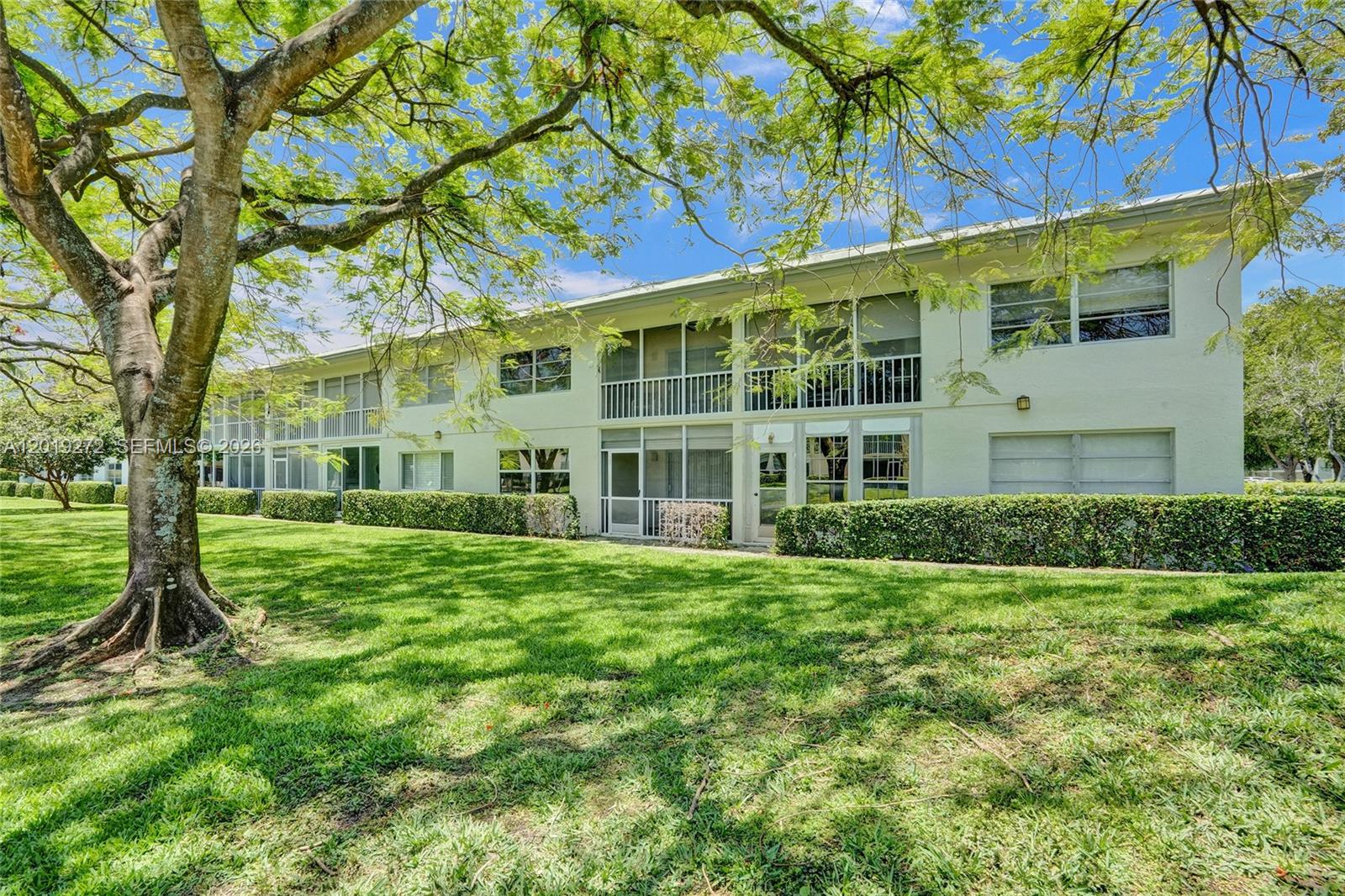 A two-story white apartment building with screened balconies sits behind a large tree and lawn — 1108 Bahama Bnd # B1, Coconut Creek FL 33066, COCONUT CREEK, FL