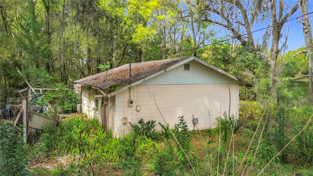 A small, weathered, light-colored house sits in a wooded area with overgrown vegetation — 10160 SE 49TH COURT, BELLEVIEW, FL