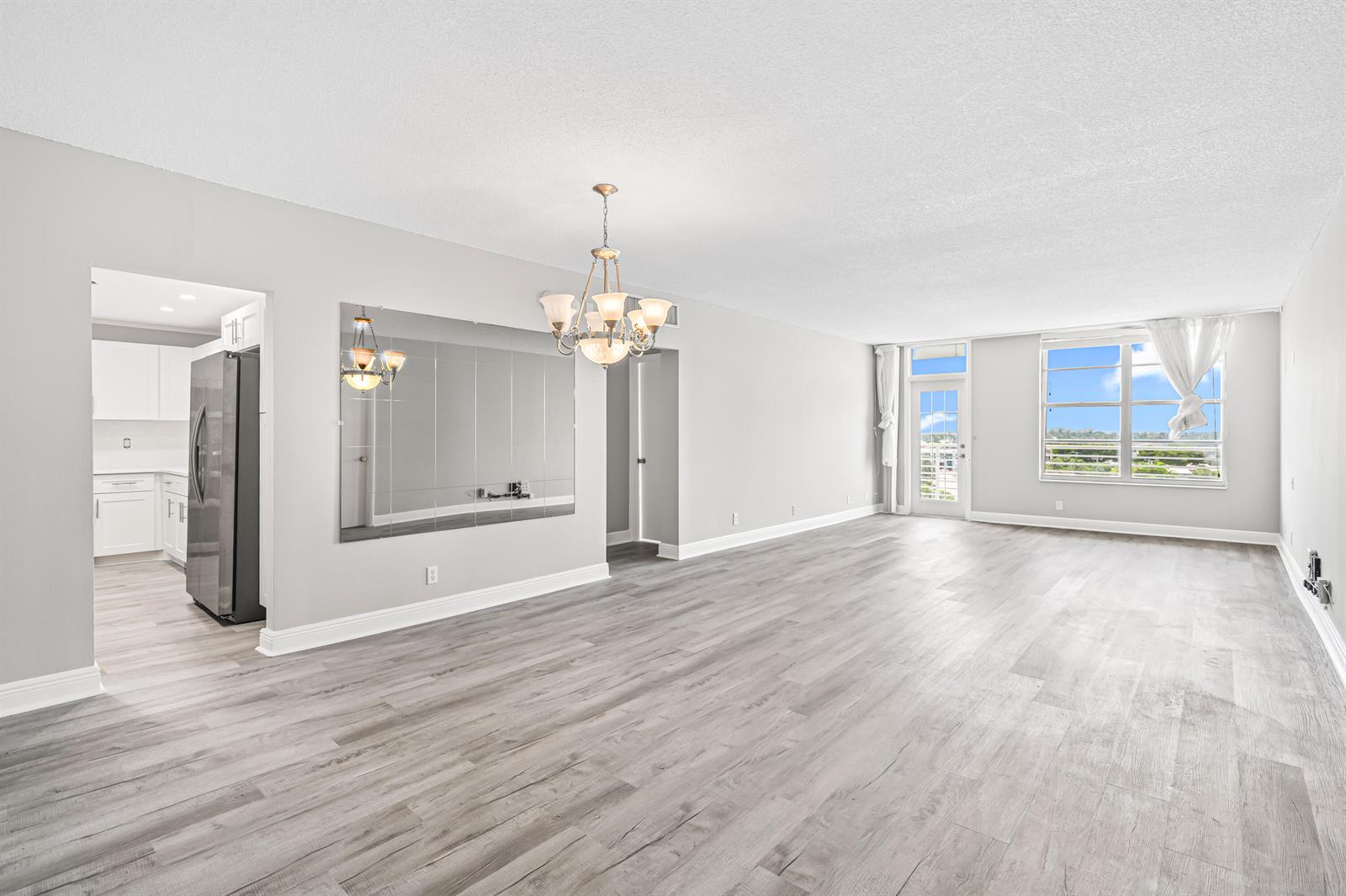 A spacious, light-filled living room features gray wood-look flooring, a large wall mirror, and chandelier — 2851 S Ocean # 0026, Boca Raton FL 33432, BOCA RATON, FL