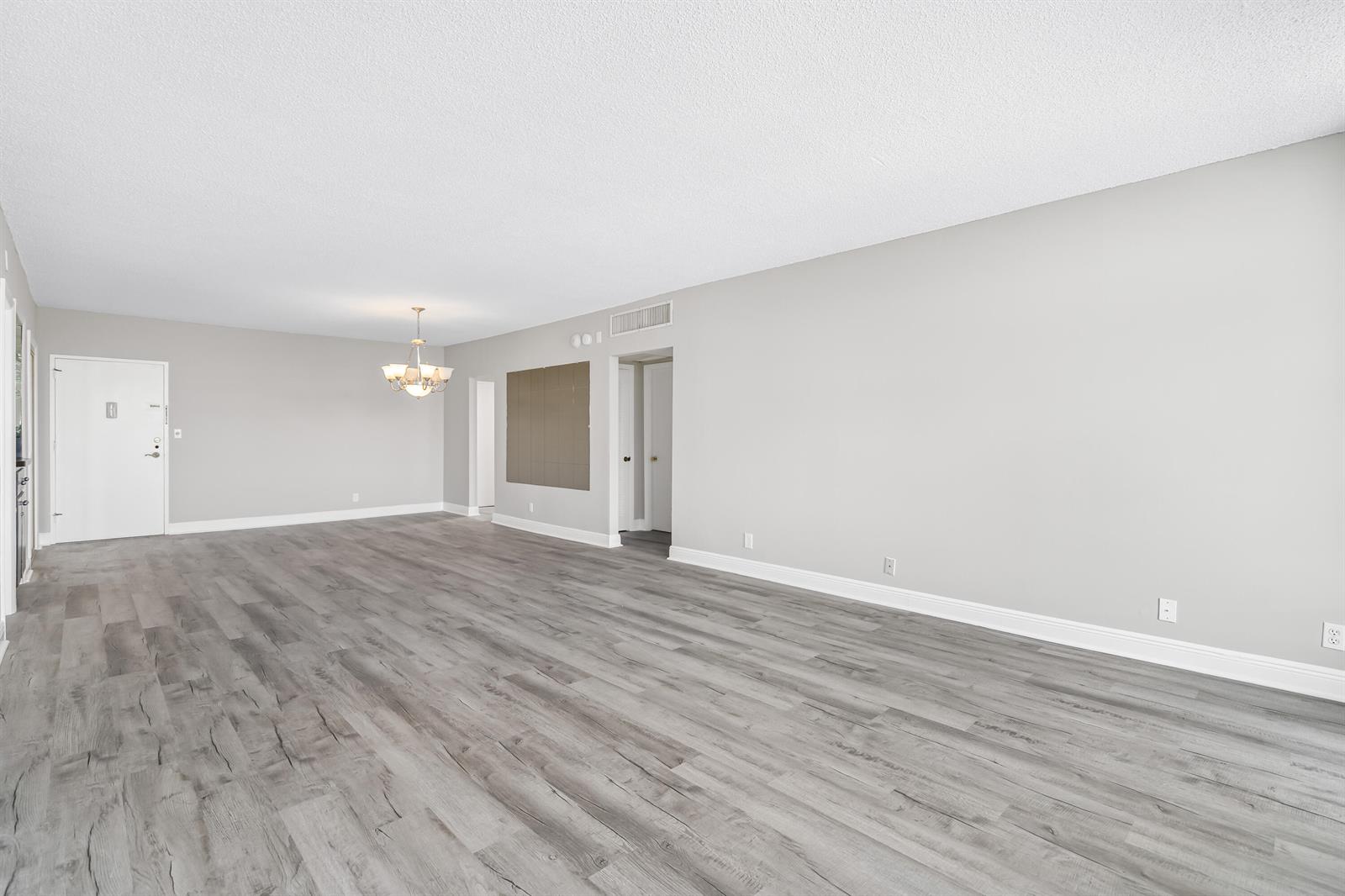 An empty living room features light gray walls, wood-look flooring, and a central chandelier — 2851 S Ocean # 0026, Boca Raton FL 33432, BOCA RATON, FL