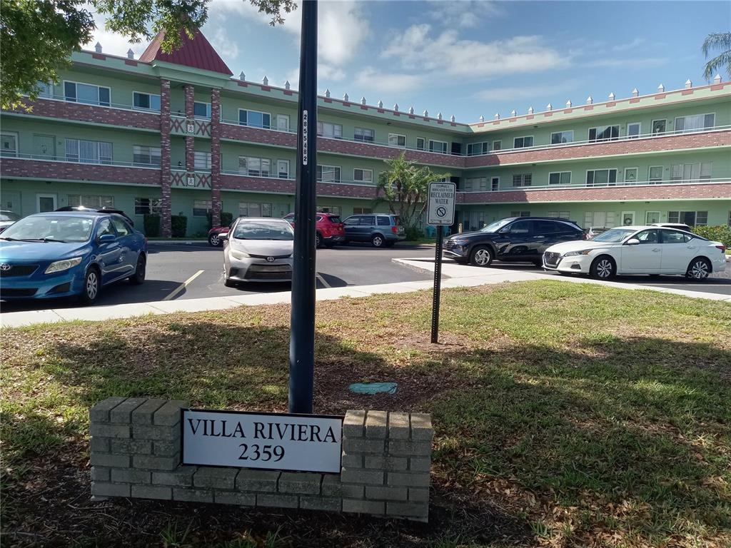 A multi-story green apartment building stands behind a parking lot with a brick sign — 2359 FINLANDIA LANE UNIT 65, CLEARWATER, FL