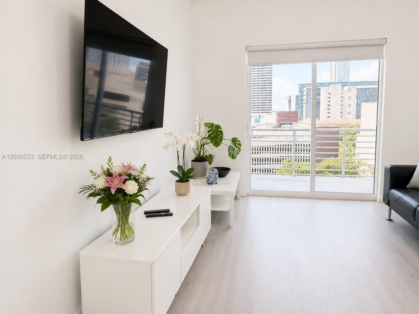 A bright living room features a wall-mounted television, white media console, and a balcony view — 234 NE 3rd St # 905, Miami FL 33132, MIAMI, FL