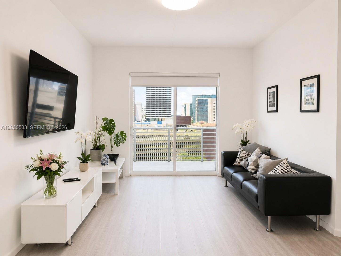 A bright living room features a black leather sofa, white media console, and balcony access — 234 NE 3rd St # 905, Miami FL 33132, MIAMI, FL