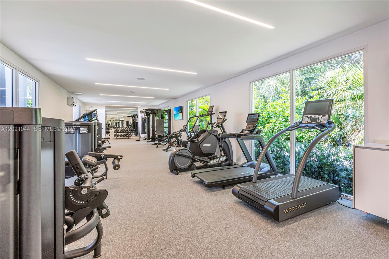 A modern gym features various exercise machines, large windows overlooking greenery, and recessed ceiling lighting — 10250 Collins Avenue # 408, Bal Harbour FL 33154, BAL HARBOUR, FL