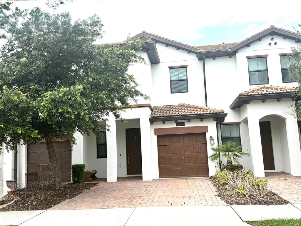A two-story townhouse exterior features white stucco walls, brown garage doors, and a paved driveway — 1344 ROYAL ST GEORGE BOULEVARD, DAVENPORT, FL