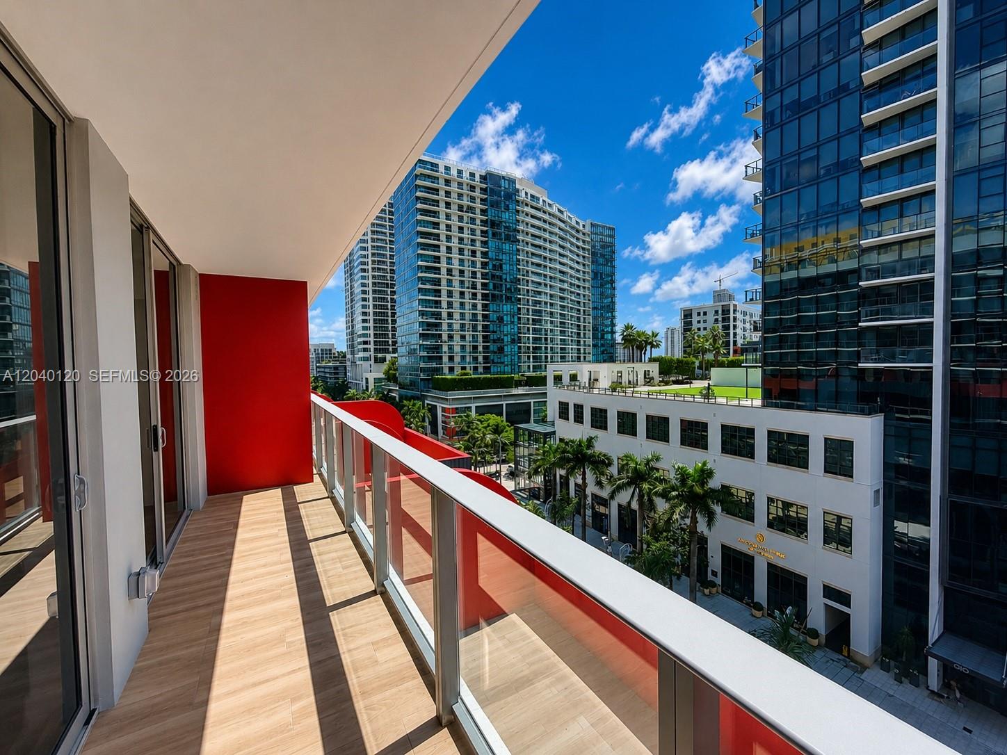A balcony with wood-look flooring, a red accent wall, and a glass railing overlooking buildings — 90 NE 32nd St # 705, Miami FL 33137, MIAMI, FL