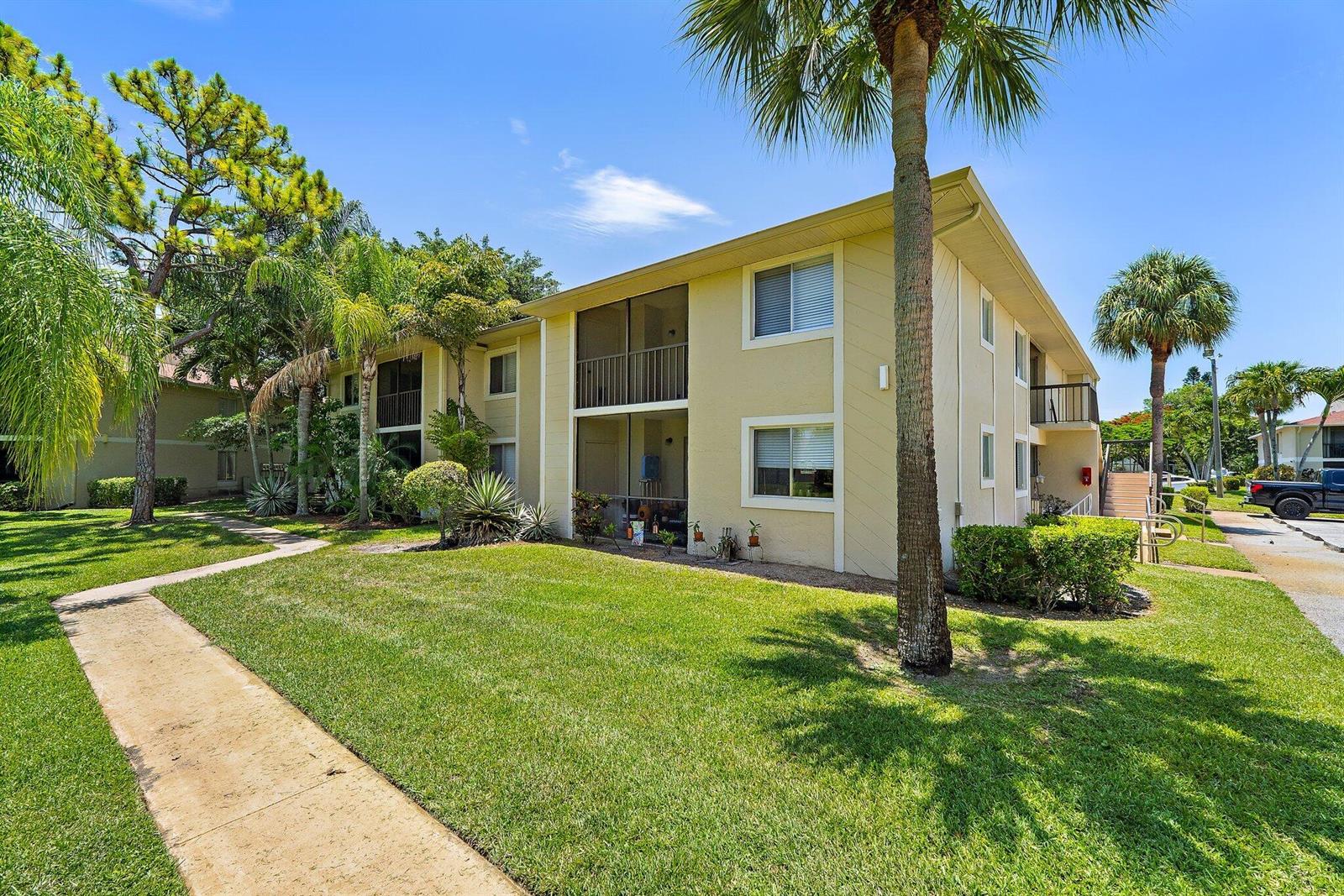 A two-story apartment building with beige siding, screened balconies, and surrounding palm trees and lawn — 6531 Chasewood Drive # H, Jupiter FL 33458, JUPITER, FL