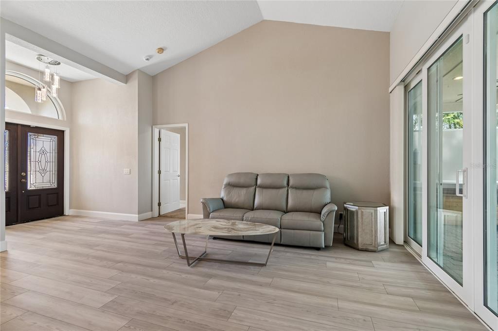A living room with vaulted ceilings, light wood-look flooring, a grey sofa, and sliding glass doors — 4227 AUSTON WAY, PALM HARBOR, FL