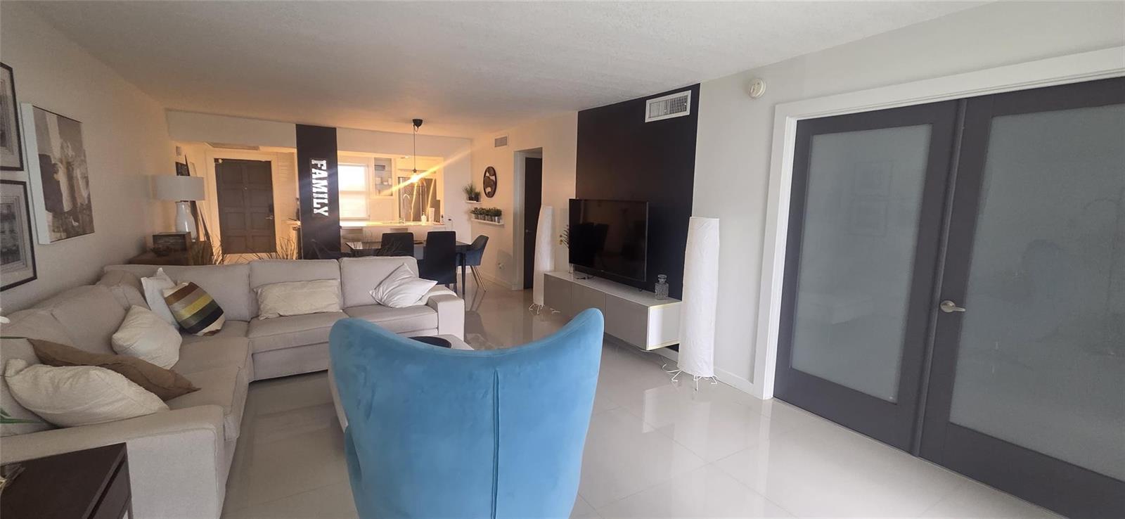 A living room features a beige sectional sofa, a blue armchair, and a wall-mounted television — 2671 S Course Drive # 806, Pompano Beach FL 33069, POMPANO BEACH, FL