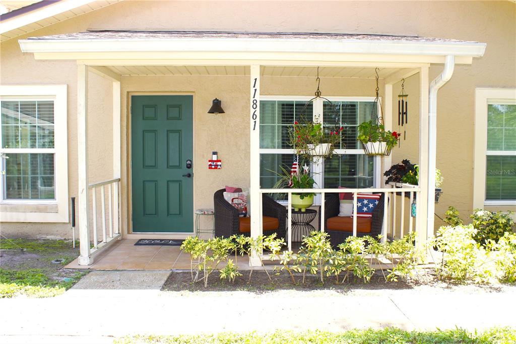 A covered front porch features a green door, two wicker chairs, and hanging potted plants — 11861 RAINTREE DRIVE UNIT 65, TAMPA, FL