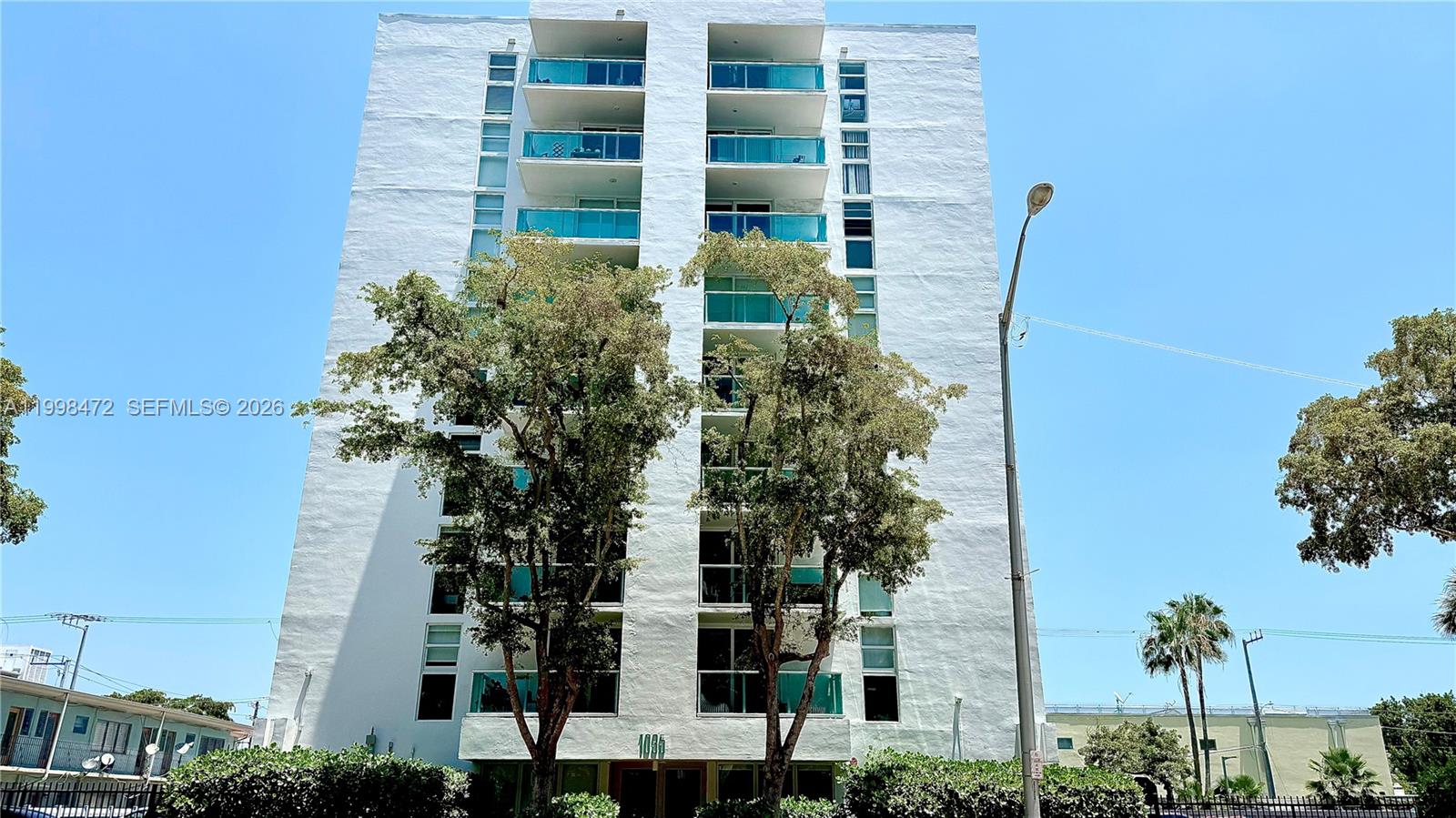 A multi-story white apartment building stands behind two large trees under a clear sky — 1035 West Ave # 306, Miami Beach FL 33139, MIAMI BEACH, FL