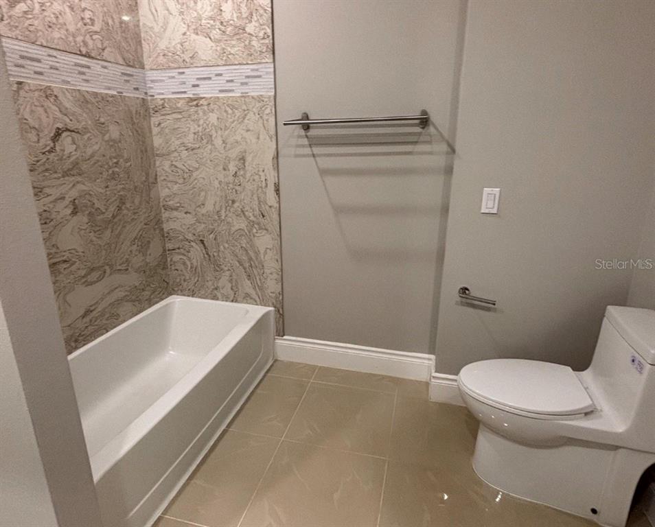 A bathroom features a white bathtub with marble-patterned tile walls, a toilet, and tile flooring — 1915 GREENBRIAR TERRACE, DAVENPORT, FL