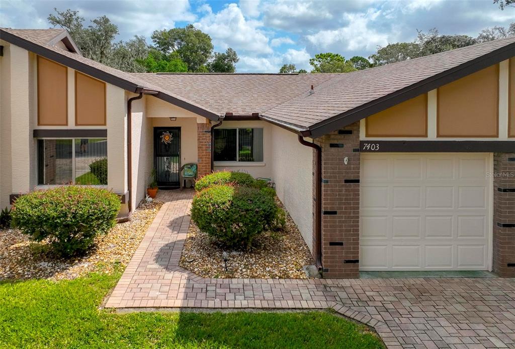 A single-story house exterior features a brick-accented garage, a paved walkway, and landscaping — 7403 HEATHER WALK DRIVE, WEEKI WACHEE, FL