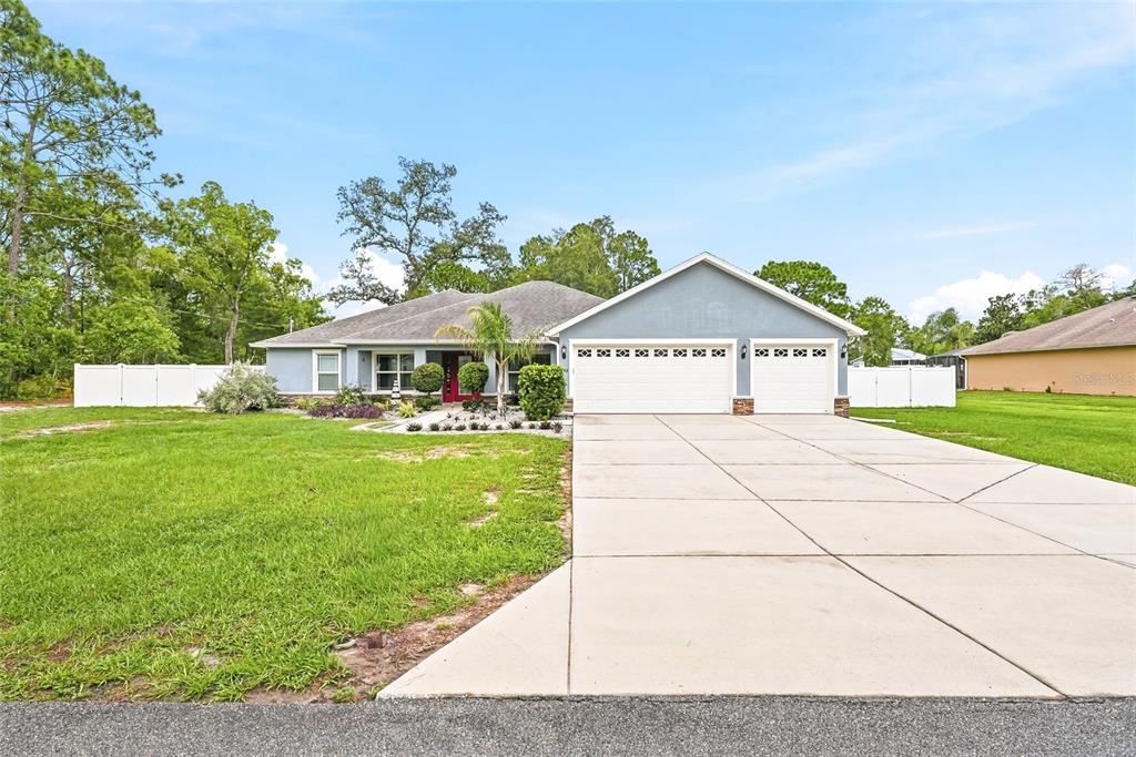 A single-story house with a three-car garage, a red front door, and a white fence — 1180 BATTERSEA AVENUE, SPRING HILL, FL