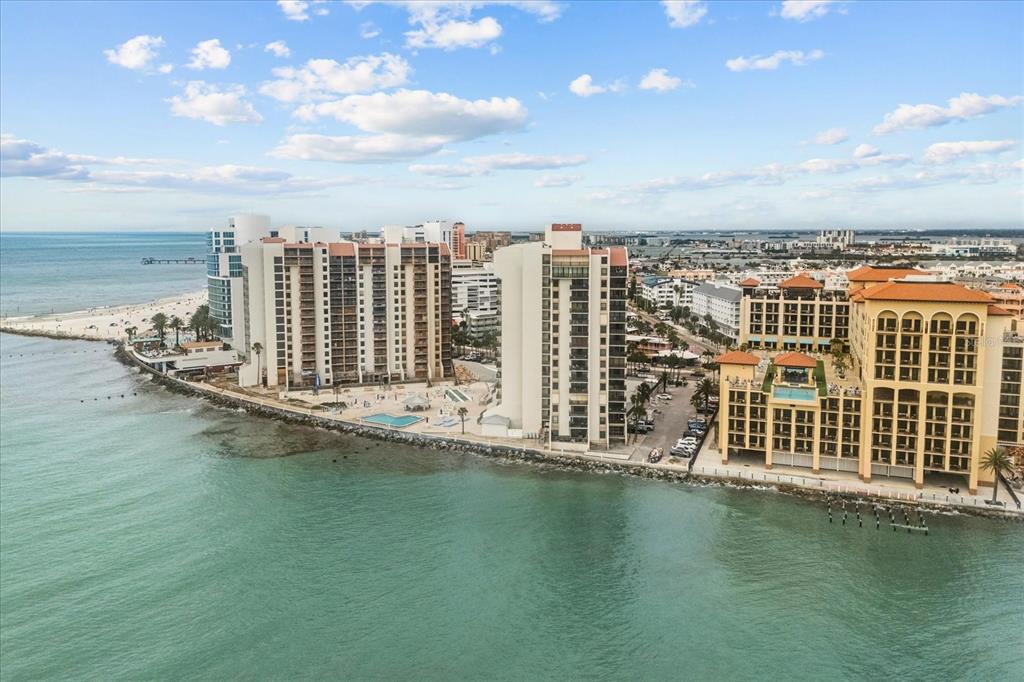 An aerial view shows high-rise beachfront condominiums along a rocky coastline under a blue sky — 450 S GULFVIEW BOULEVARD UNIT 802, CLEARWATER BEACH, FL