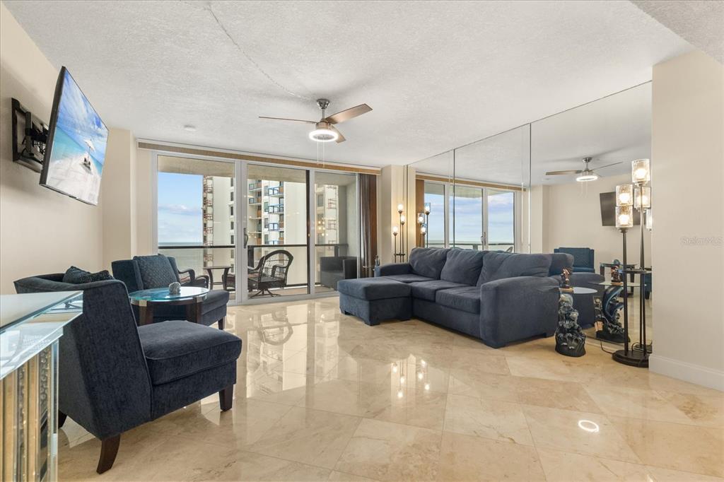 A living room features a blue sectional sofa, matching armchairs, tile flooring, and mirrored walls — 450 S GULFVIEW BOULEVARD UNIT 802, CLEARWATER BEACH, FL