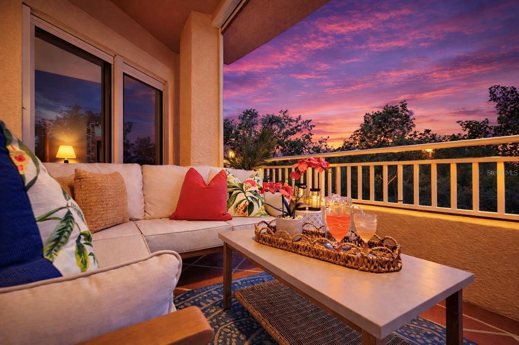 A furnished balcony features a sofa, coffee table, and sunset views over the trees — 12033 GANDY BOULEVARD N UNIT 133, ST PETERSBURG, FL