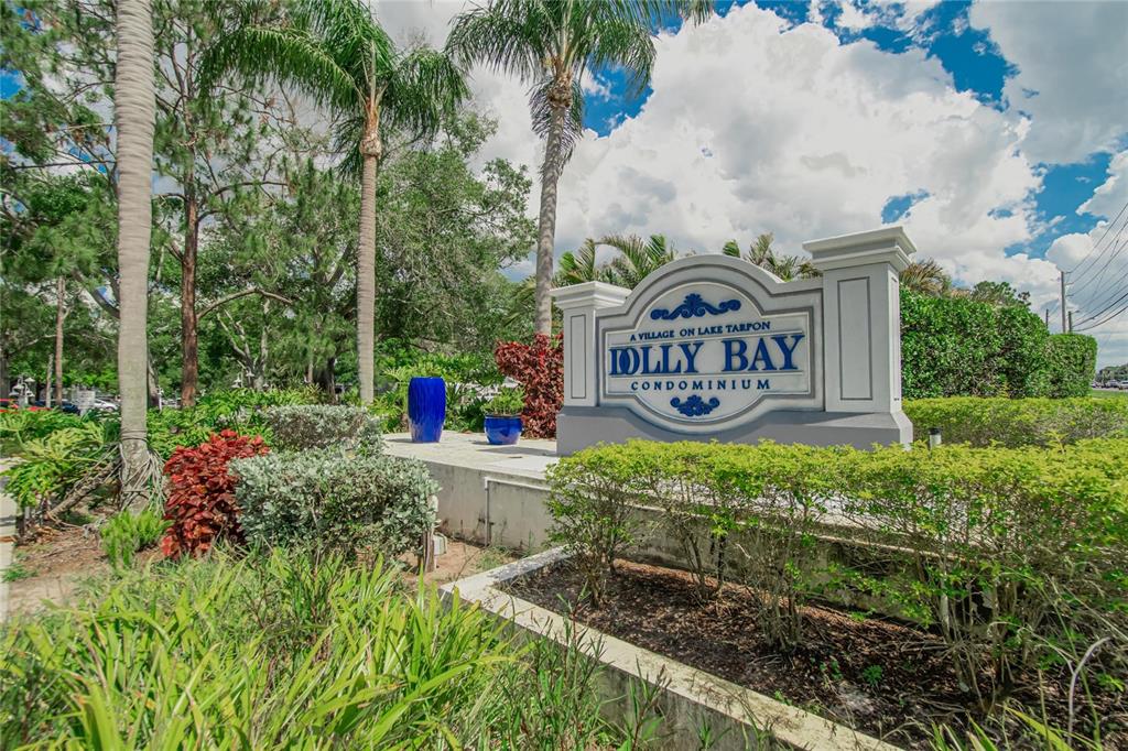 The entrance sign for Jolly Bay Condominium stands among palm trees and landscaped garden beds — 2599 DOLLY BAY DRIVE UNIT 305, PALM HARBOR, FL