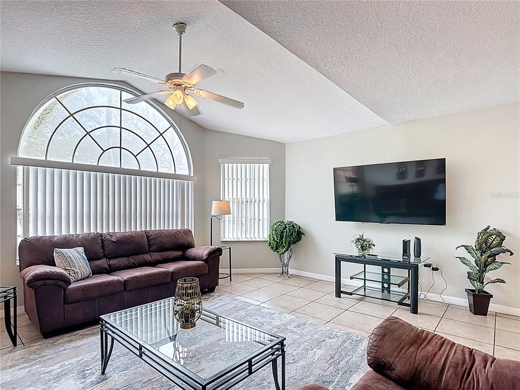 A living room features a large arched window, tile flooring, a ceiling fan, and a wall-mounted television — 8710 RAIN FOREST PLACE UNIT B, KISSIMMEE, FL