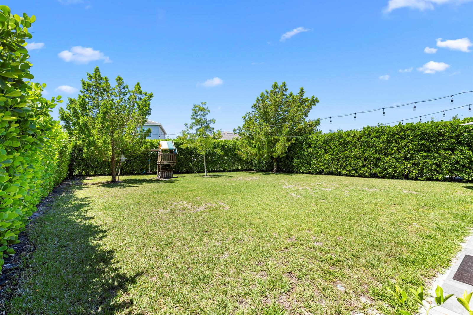 A grassy backyard features a small wooden play structure, mature trees, and a tall hedge — 16201 Melogold Drive, Westlake FL 33470, WESTLAKE, FL