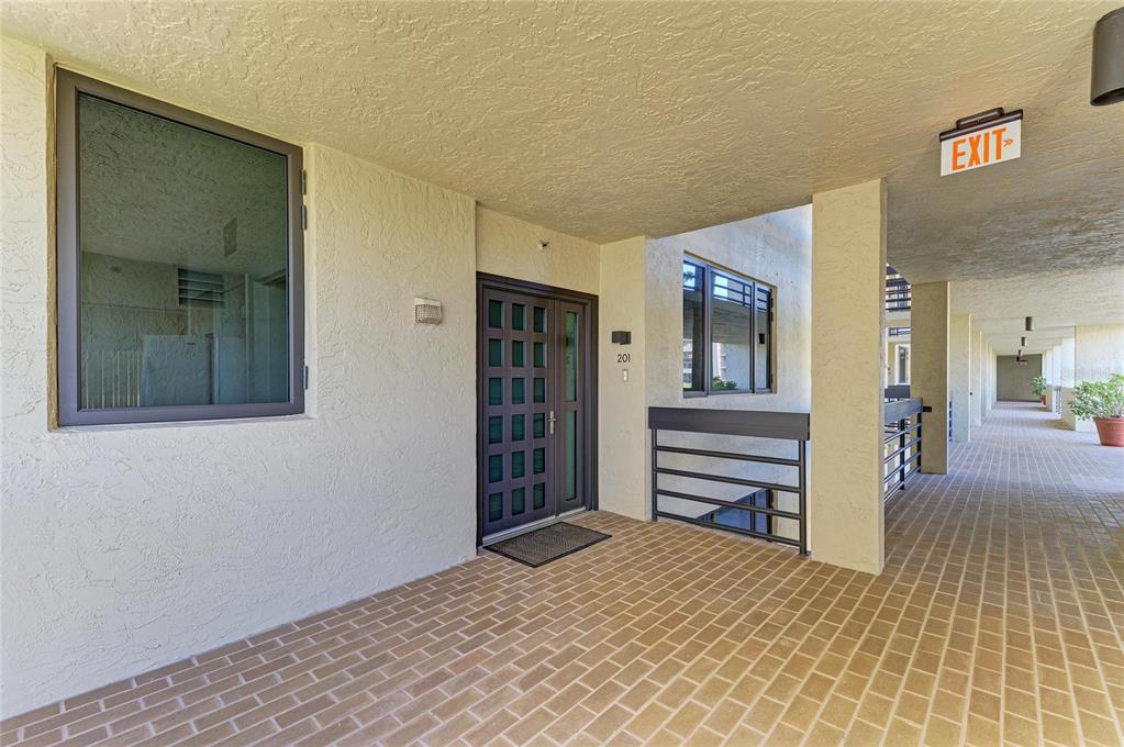 An outdoor apartment hallway features a brick floor, a dark door, and an exit sign — 1065 GULF OF MEXICO DRIVE UNIT 201, LONGBOAT KEY, FL