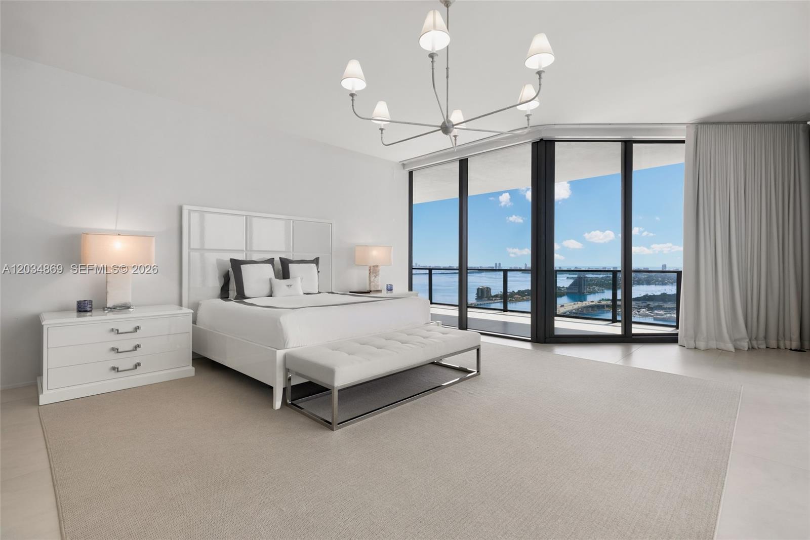 A modern bedroom features a white bed, a chandelier, and floor-to-ceiling windows with water views — 1000 Biscayne Blvd # 3701, Miami FL 33132, MIAMI, FL