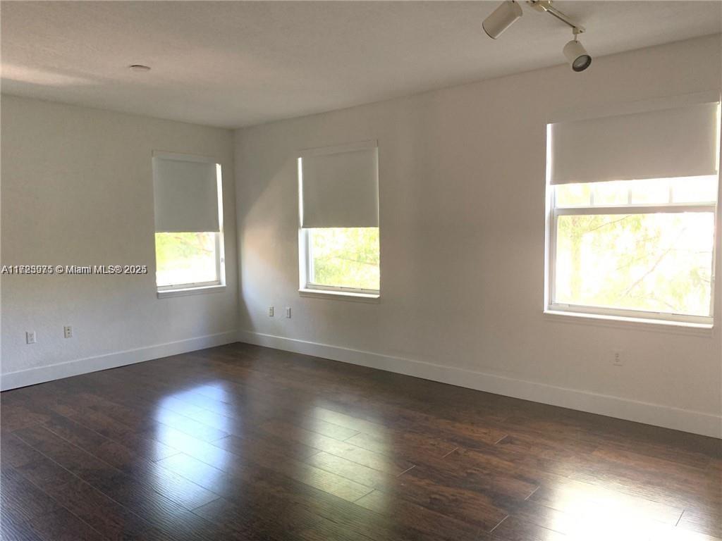 An empty room features dark wood flooring, white walls, and three windows with white shades — 6776 W Sample Rd # 6776, Coral Springs FL 33067, CORAL SPRINGS, FL