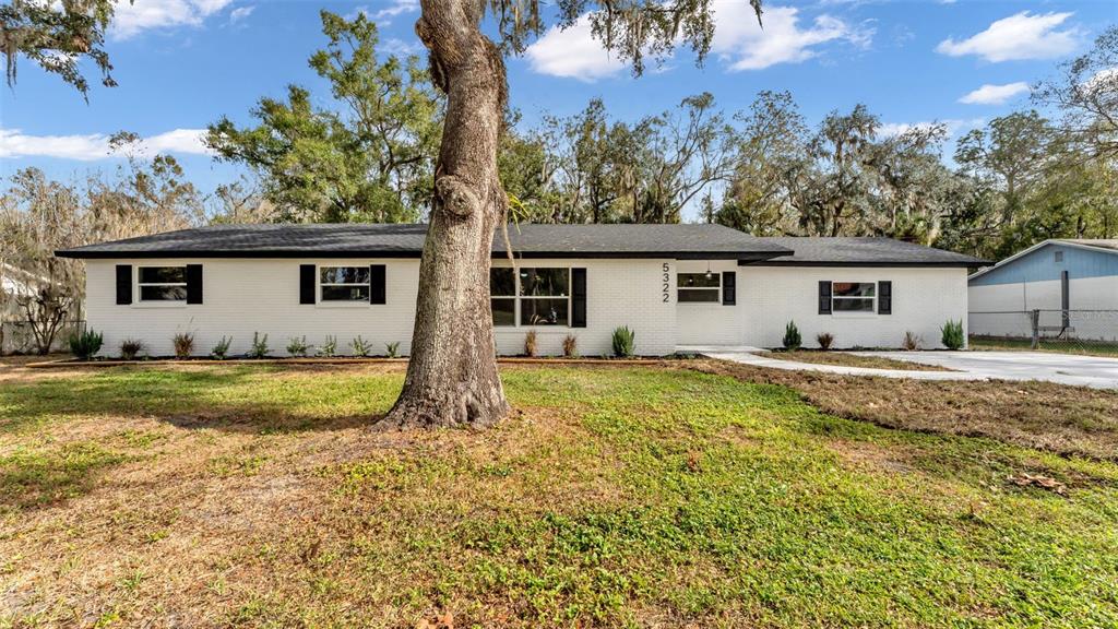 A single-story white brick house with a dark roof and black shutters sits behind a tree — 5322 FORESTBROOK DRIVE E, LAKELAND, FL