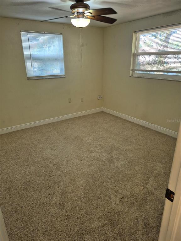 An empty bedroom features beige walls, tan carpeting, two windows with blinds, and a ceiling fan — 6616 HONE STREET, NEW PORT RICHEY, FL