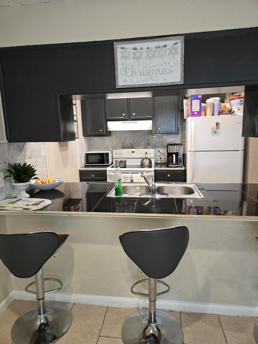 A kitchen features dark cabinetry, black countertops, a breakfast bar with stools, and white appliances — 2396 Daphne Drive, West Palm Beach FL 33415, WEST PALM BEACH, FL