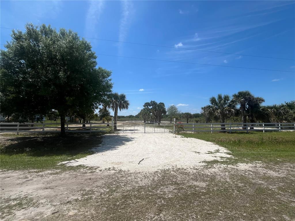 A gravel driveway leads to a gated entrance with a white fence on a sunny day — 19374 NW 302ND STREET, OKEECHOBEE, FL