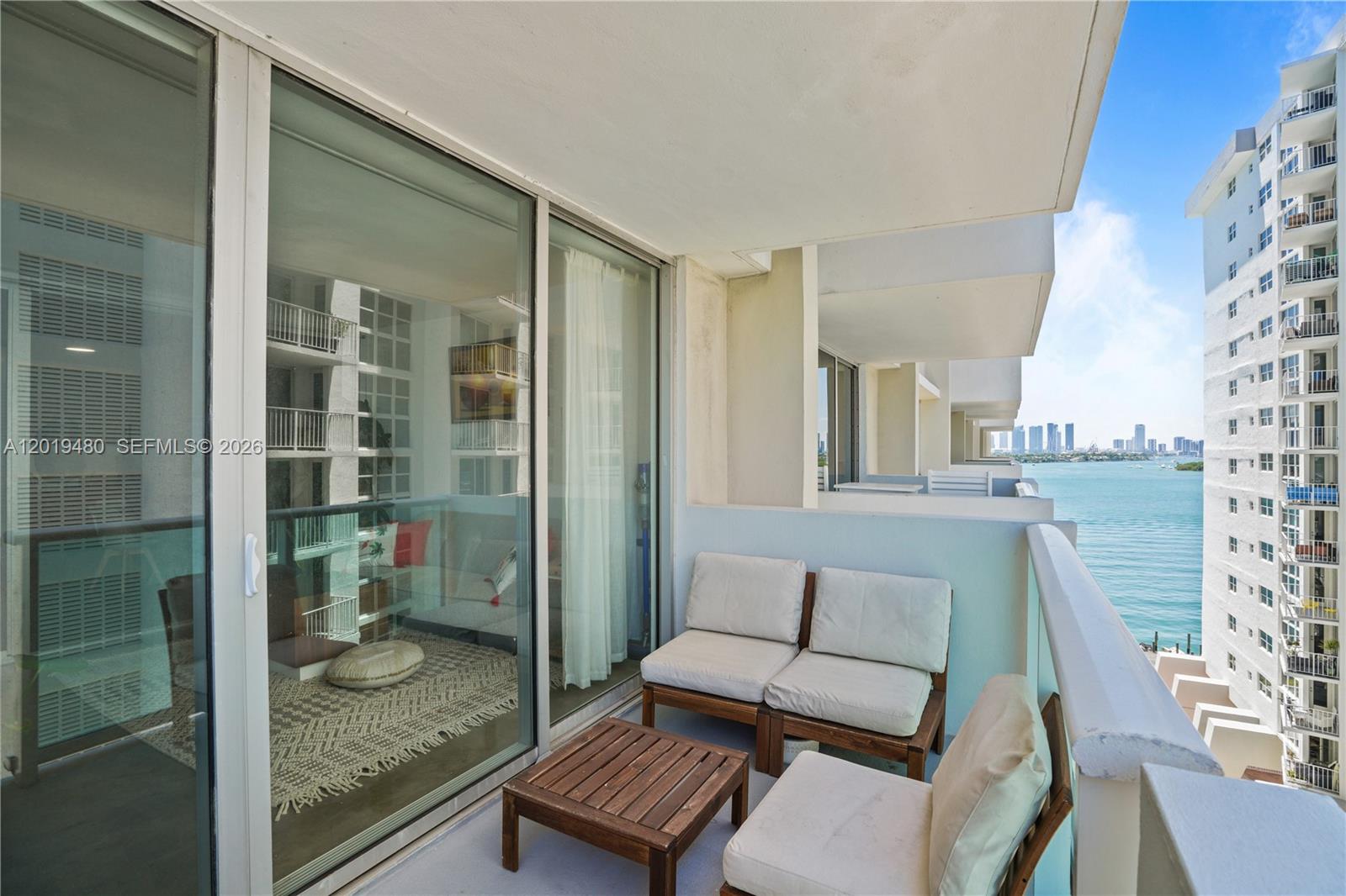 A balcony with outdoor seating overlooks a body of water and a neighboring high-rise building — 1200 West Ave # 819, Miami Beach FL 33139, MIAMI BEACH, FL