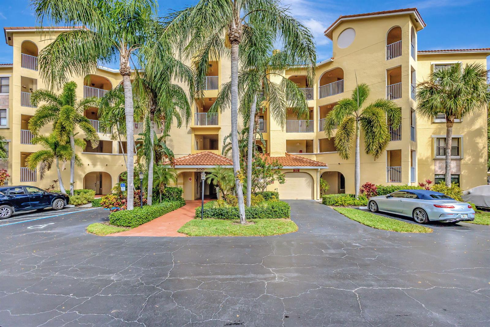 A multi-story yellow apartment building with palm trees, a parking lot, and a covered entrance — 300 Uno Lago Drive # 101, Juno Beach FL 33408, JUNO BEACH, FL