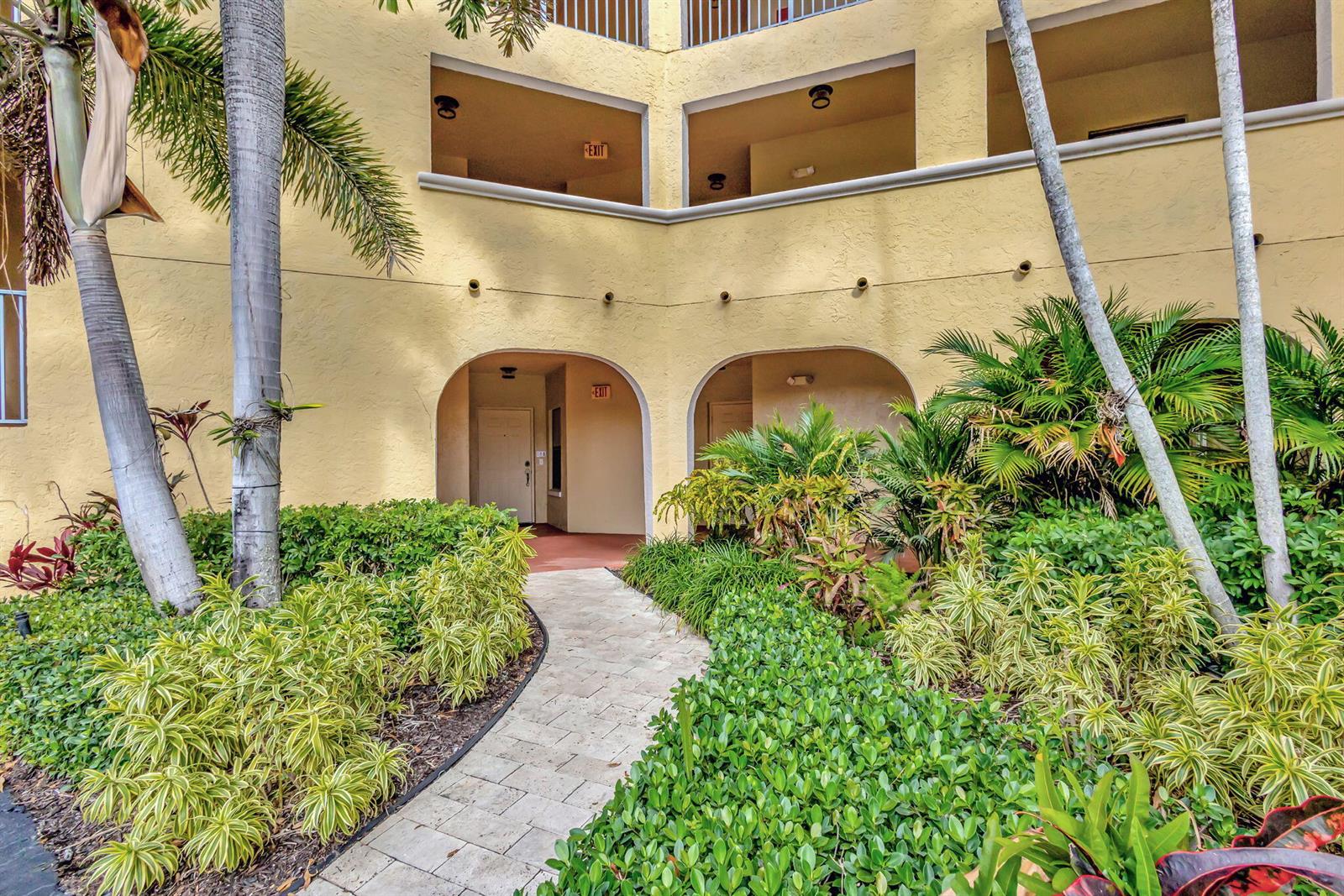 A stone walkway leads to two arched entrances of a yellow multi-story building with landscaping — 300 Uno Lago Drive # 101, Juno Beach FL 33408, JUNO BEACH, FL