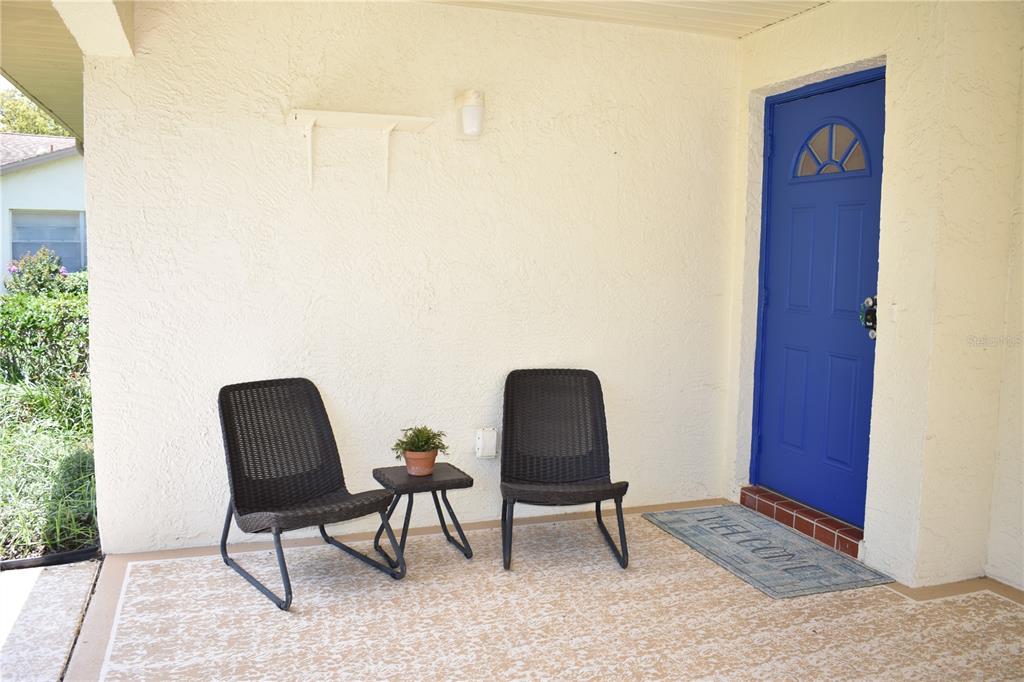 A covered porch features two dark wicker chairs, a small side table, and a blue door — 2005 HALMROCK PLACE UNIT 174, SUN CITY CENTER, FL