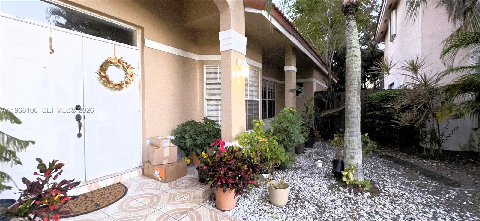 The exterior of a house features a covered entryway with potted plants and stacked packages — 5769 NW 99th Ave, Doral FL 33178, DORAL, FL