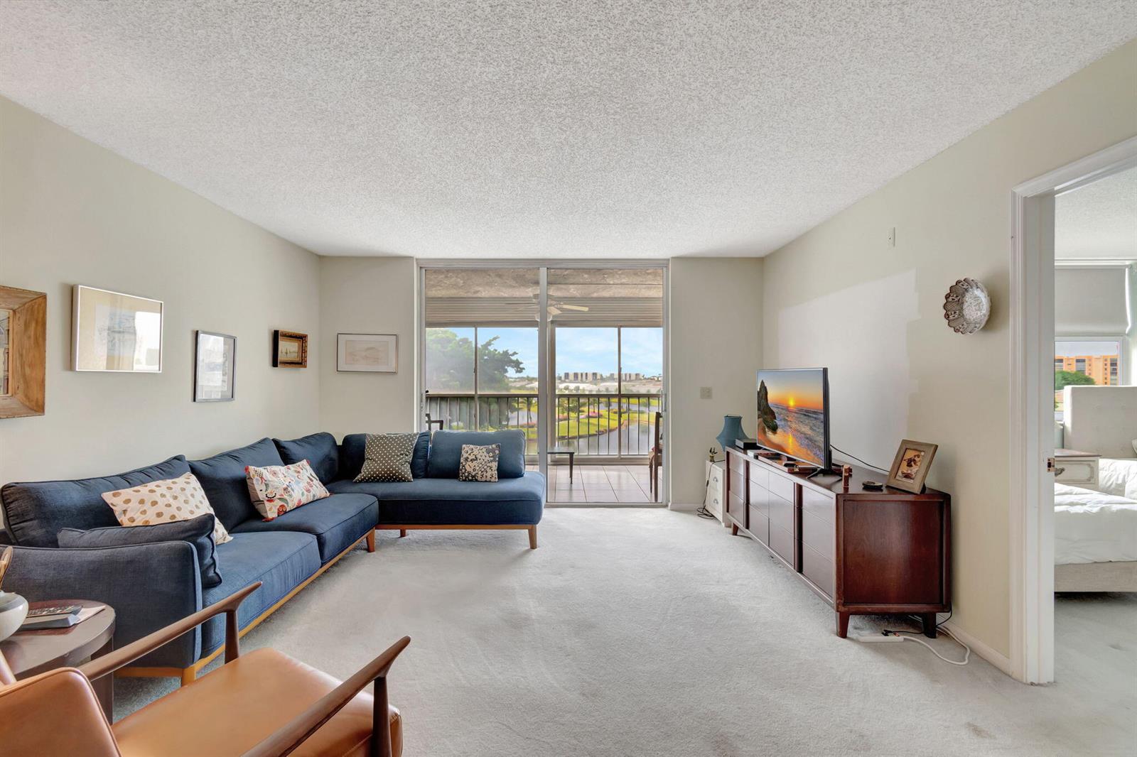 A living room with a blue sectional sofa, a wooden media console, and a balcony view — 14575 Bonaire Boulevard # 402, Delray Beach FL 33446, DELRAY BEACH, FL