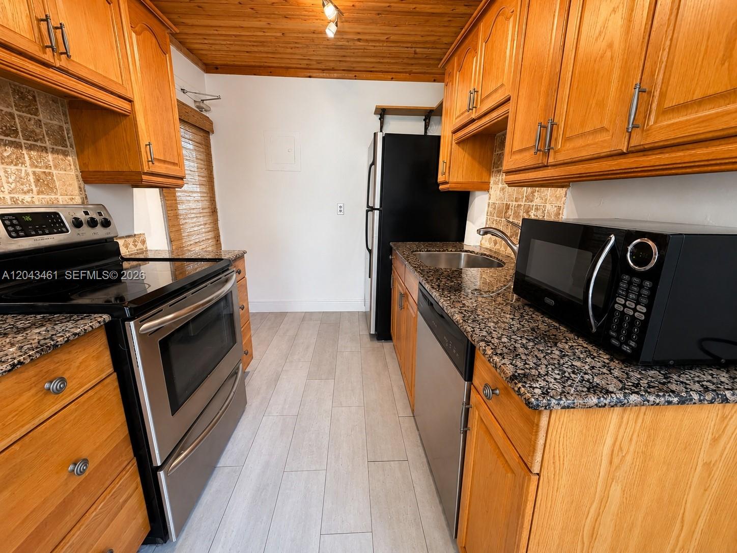 A galley kitchen features oak cabinets, granite countertops, stainless steel appliances, and wood-paneled ceilings — 1670 Bay Rd # 3D, Miami Beach FL 33139, MIAMI BEACH, FL
