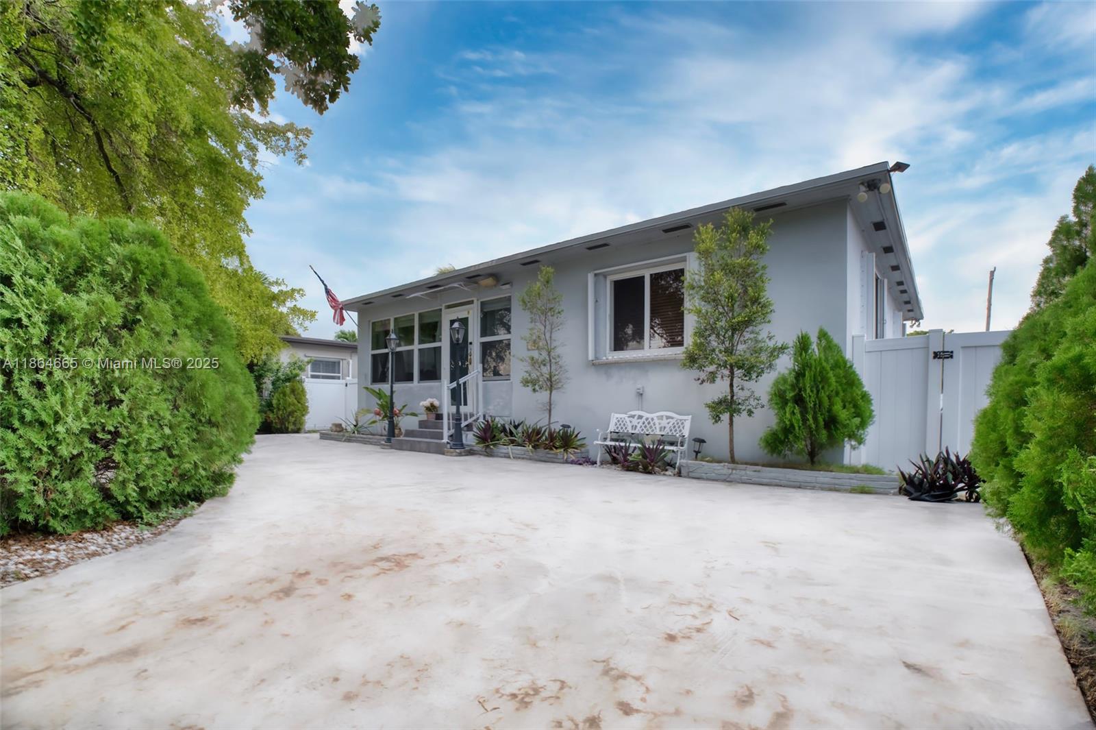 A single-story gray house features a concrete driveway, a white bench, and surrounding landscaping — 1923 Dewey St, Hollywood FL 33020, HOLLYWOOD, FL
