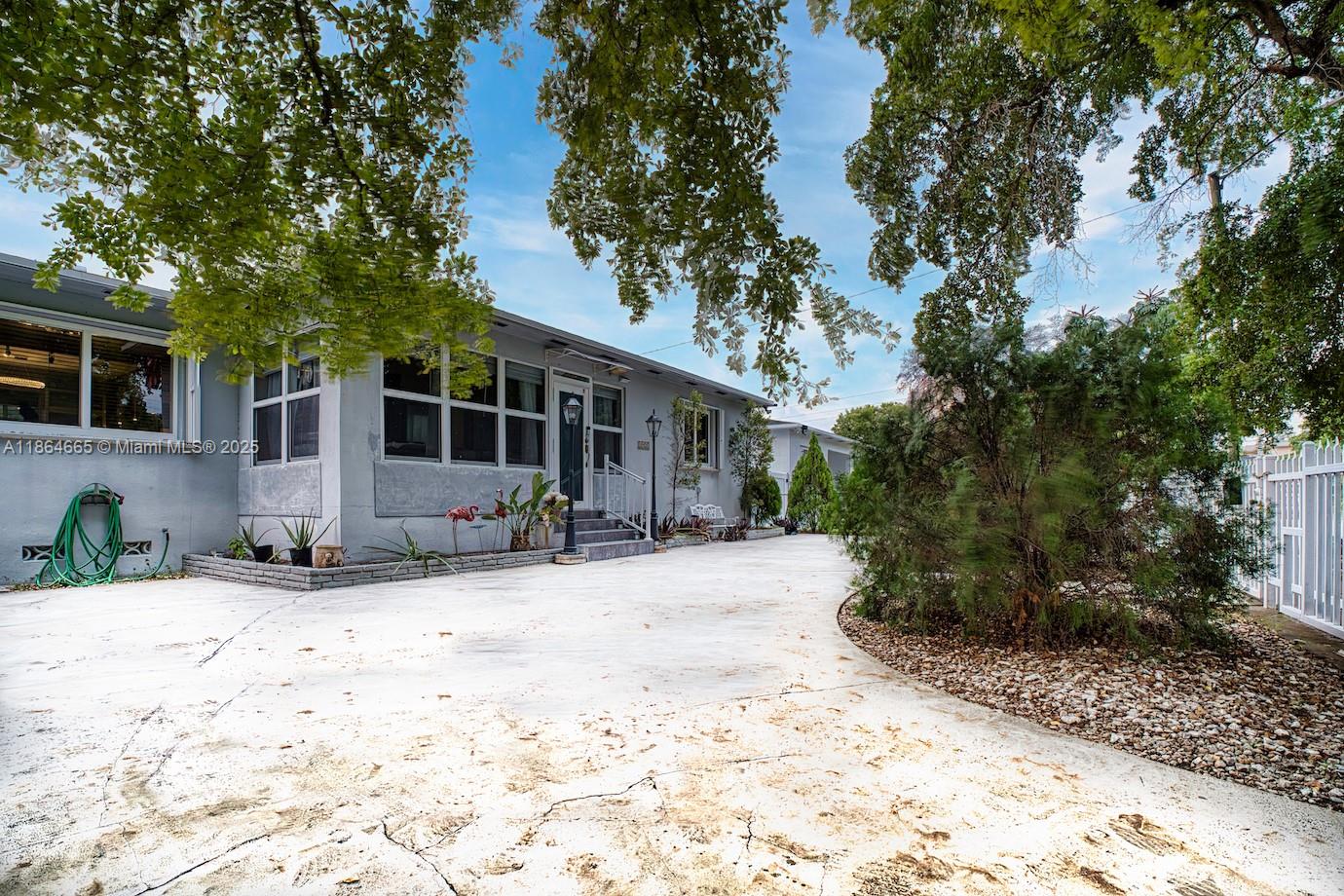 A single-story gray house features a concrete driveway, large trees, and a white fence — 1923 Dewey St, Hollywood FL 33020, HOLLYWOOD, FL