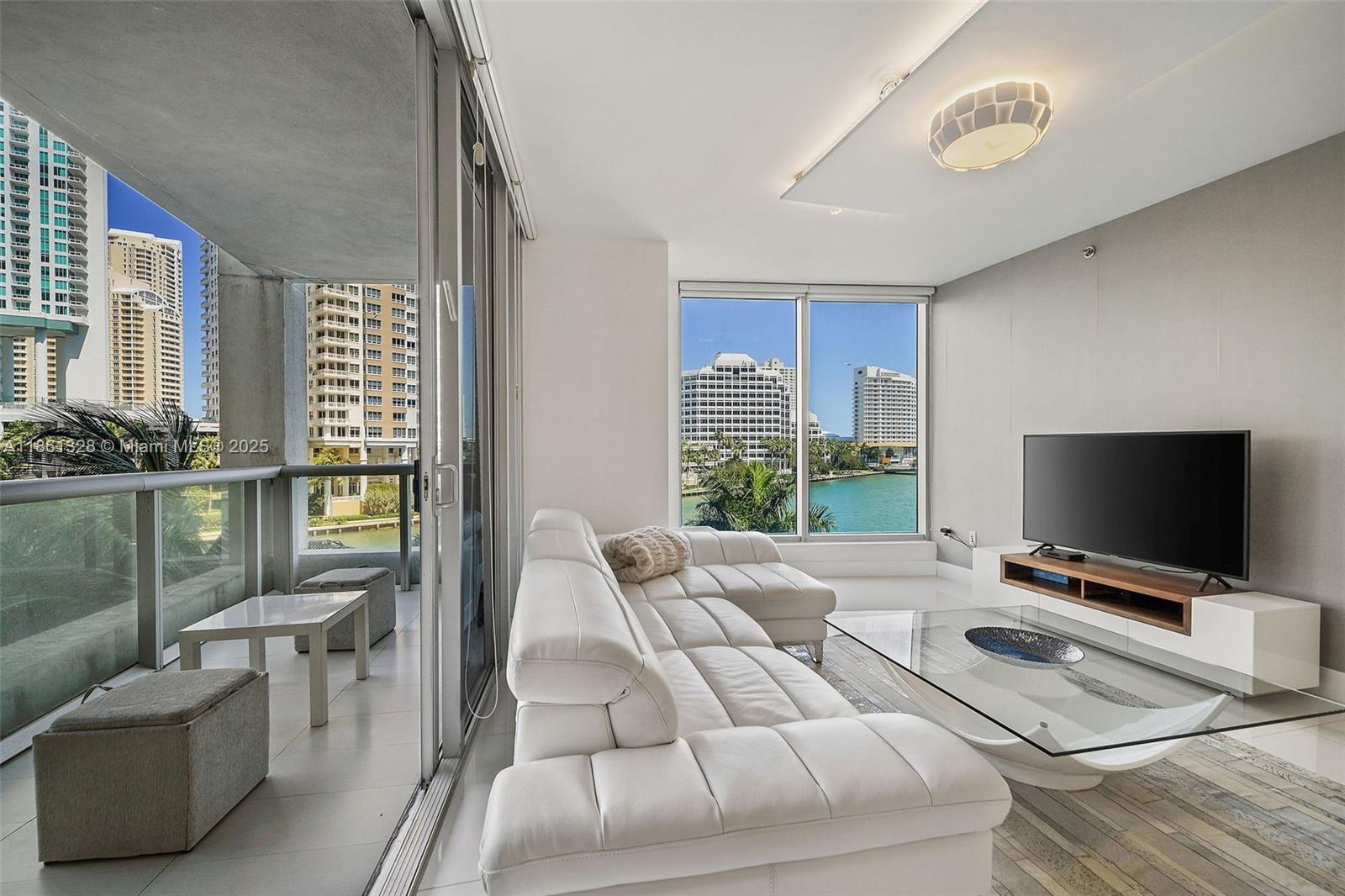 A modern living room features a white sectional sofa, glass coffee table, and balcony overlooking water — 495 Brickell Ave # 402, Miami FL 33131, MIAMI, FL