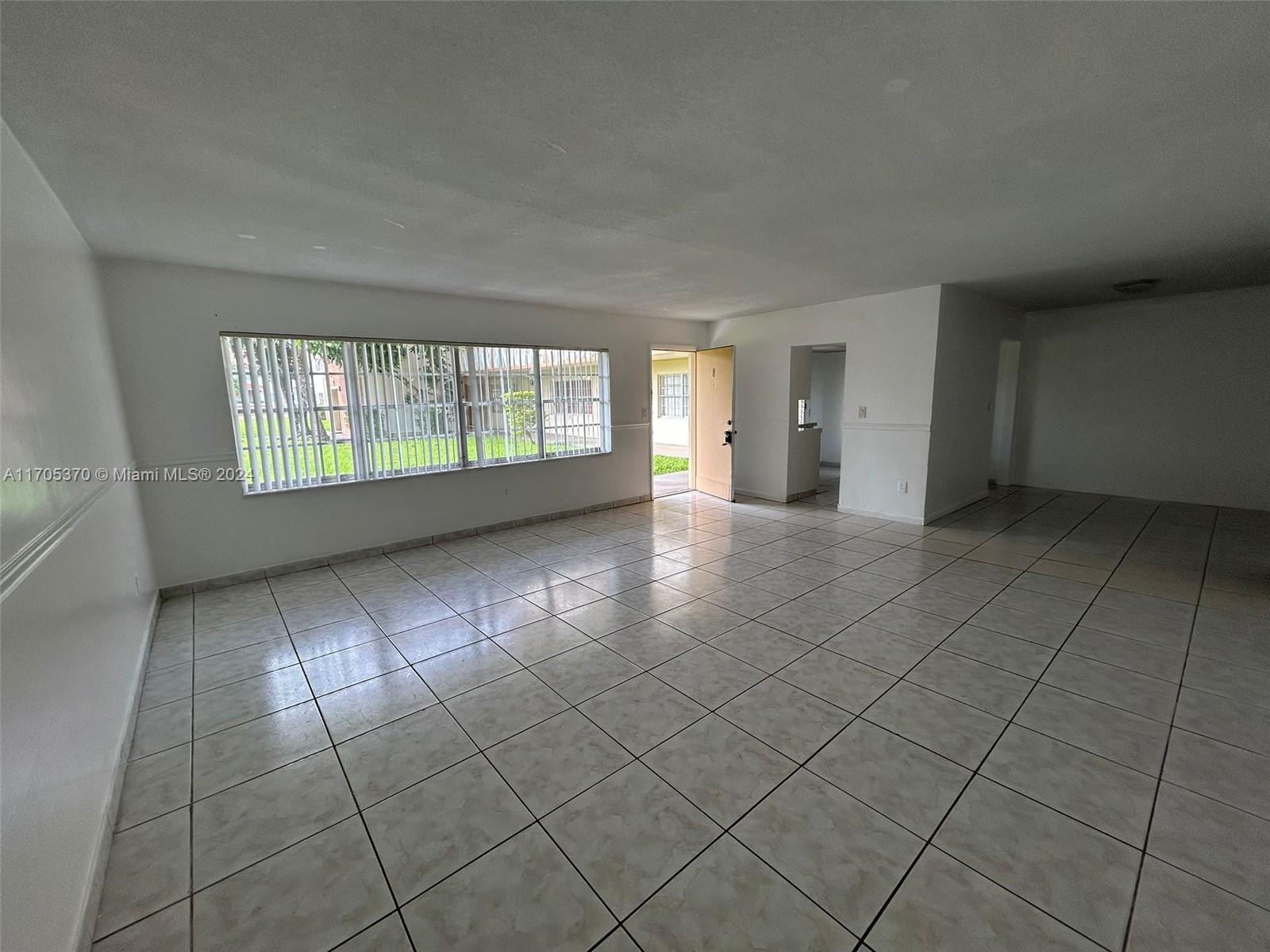 An empty living room features white walls, light-colored tile flooring, and a large window — 11855 NE 19th Dr # 8, North Miami FL 33181, NORTH MIAMI, FL